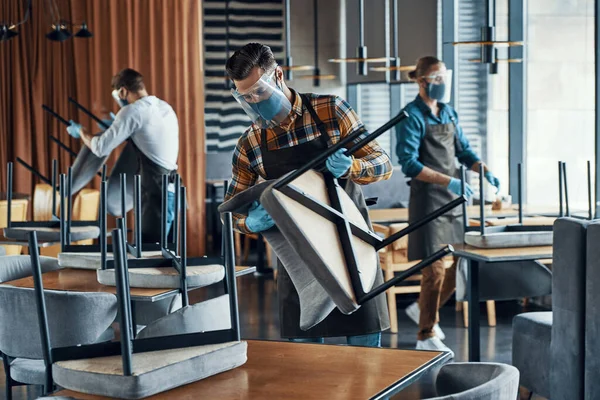 Three men in protective face shields and aprons arranging furniture in restaurant — Stock Photo, Image
