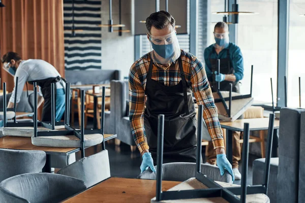 Three male waiters in protective workwear arranging furniture in restaurant — Stock Photo, Image