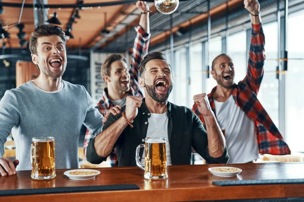 Animando os jovens em roupas casuais bebendo cerveja e assistindo a jogos esportivos enquanto sentados no pub — Fotografia de Stock
