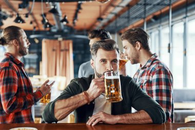 Charming young man in casual clothing drinking beer while spending time with friends in the pub