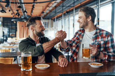 Cheerful young men in casual clothing shaking hands and drinking beer while spending time in the pub