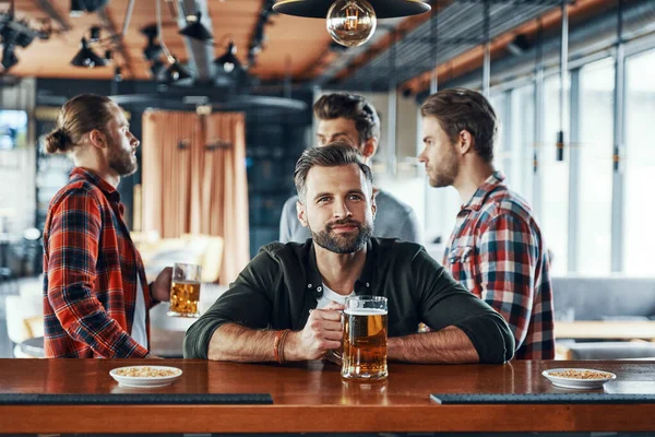 Guapo joven con ropa casual bebiendo cerveza mientras pasa tiempo con amigos en el pub —  Fotos de Stock