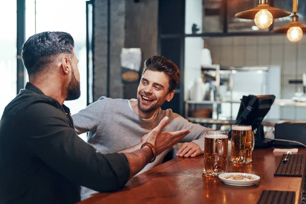 Two happy young men in casual clothing talking and drinking beer while spending time in the pub — Φωτογραφία Αρχείου