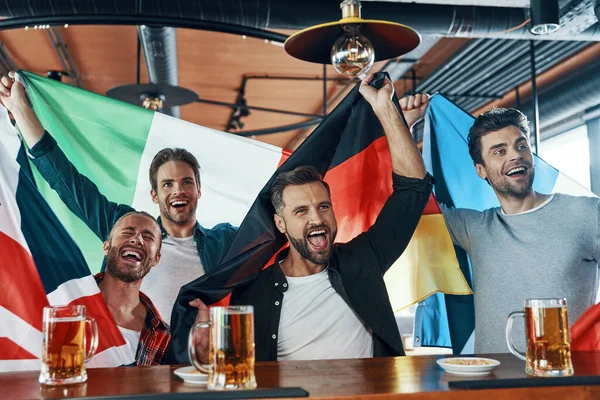 Fröhliche junge Männer mit internationalen Fahnen, die Bier trinken und sich im Pub Sportspiele anschauen — Stockfoto