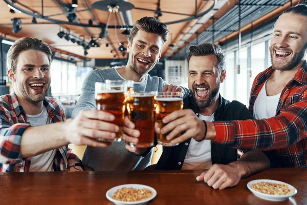 Jóvenes felices con ropa casual brindando entre sí con cerveza y riéndose mientras están sentados en el pub —  Fotos de Stock