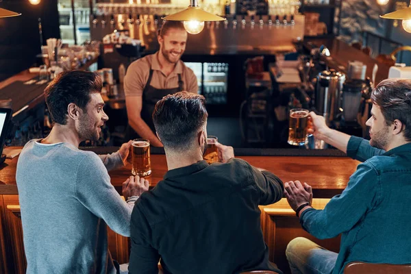 Smiling young men in casual clothing drinking beer while sitting in the pub together —  Fotos de Stock