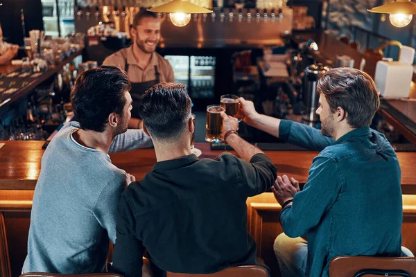 Carefree young men in casual clothing drinking beer while sitting in the pub together — Stok fotoğraf