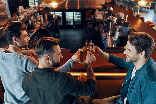 Draufsicht auf glückliche junge Männer in lässiger Kleidung, die sich im Pub mit Bier anstoßen und lächeln — Stockfoto