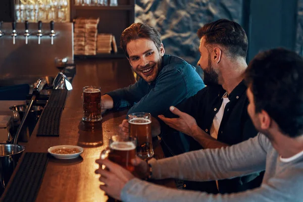 Happy young men in casual clothing drinking beer and bonding together while sitting in the pub — Foto de Stock