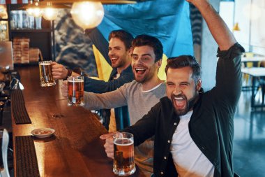 Cheering young men covered in Ukrainian flag drinking beer and watching sport game while sitting in the pub