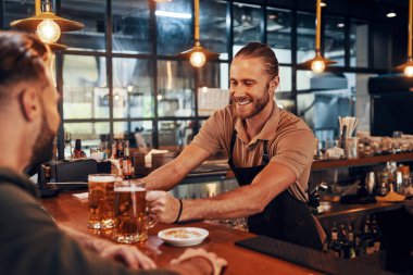 Charming young bartender in apron serving beer and smiling while working in the pub