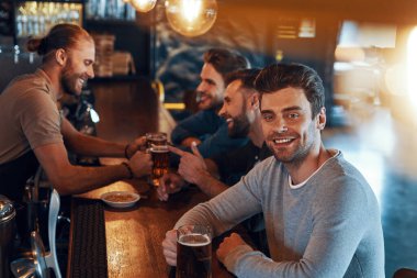 Smiling young men in casual clothing drinking beer and bonding together while sitting in the pub