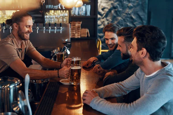 Smiling young men in casual clothing drinking beer and bonding together while sitting in the pub —  Fotos de Stock