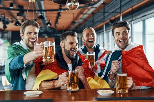 Fröhliche junge Männer mit internationalen Fahnen, die Bier trinken und sich im Pub Sportspiele anschauen — Stockfoto