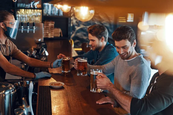 Smiling young men in casual clothing drinking beer and bonding together while sitting in the pub — ストック写真