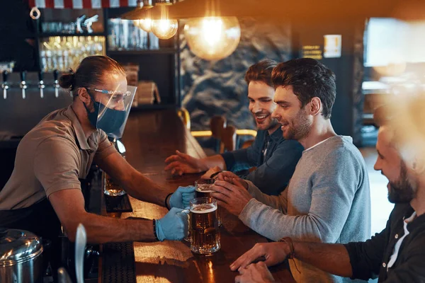 Smiling young men in casual clothing drinking beer and bonding together while sitting in the pub —  Fotos de Stock