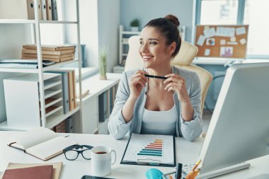 Beautiful young woman in smart casual wear working with charts and smiling while sitting in the office