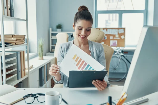 Beautiful young woman in smart casual wear working with charts and smiling while sitting in the office — Foto de Stock
