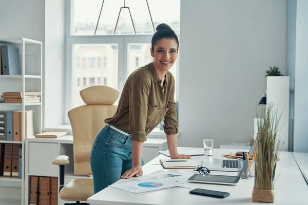 Hermosa mujer joven en ropa casual inteligente mirando a la cámara y sonriendo mientras está de pie en la oficina — Foto de Stock