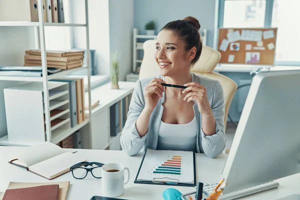 Beautiful young woman in smart casual wear working with charts and smiling while sitting in the office — Stockfoto