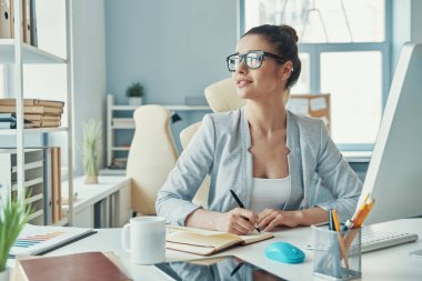 Confident young woman in smart casual wear writing something and smiling while sitting in the office