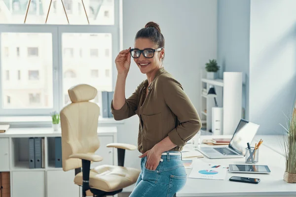 Красивая молодая женщина в элегантной повседневной одежде улыбается и регулирует очки, стоя в офисе — стоковое фото