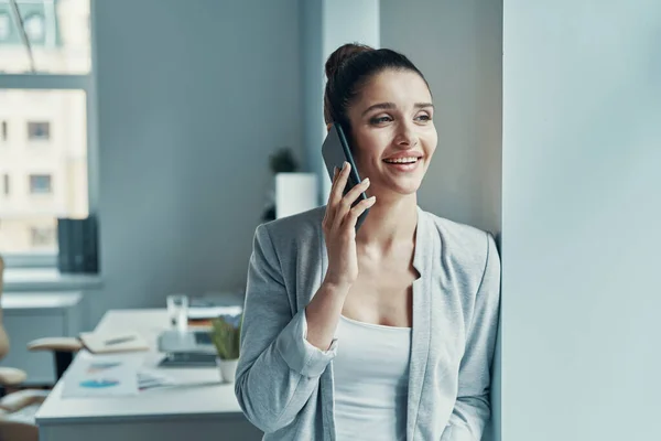 Красивая молодая женщина в умный случайный разговор по телефону и улыбаясь, стоя в офисе — стоковое фото