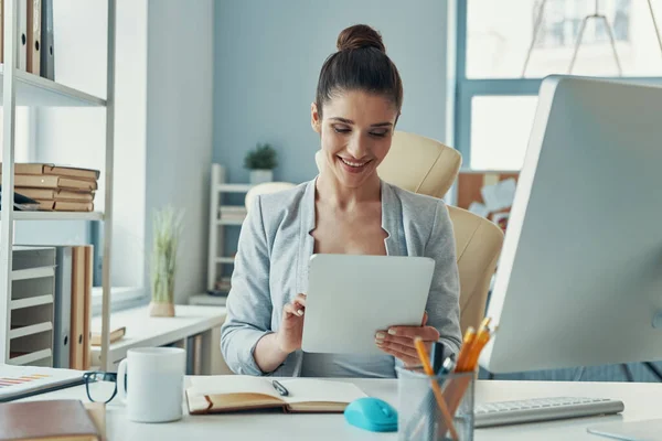 Beautiful young woman in smart casual wear using digital tablet and smiling while sitting in the office — Zdjęcie stockowe