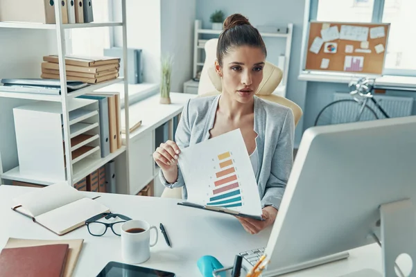 Thoughtful young woman in smart casual wear working with charts while sitting in the office — Fotografia de Stock