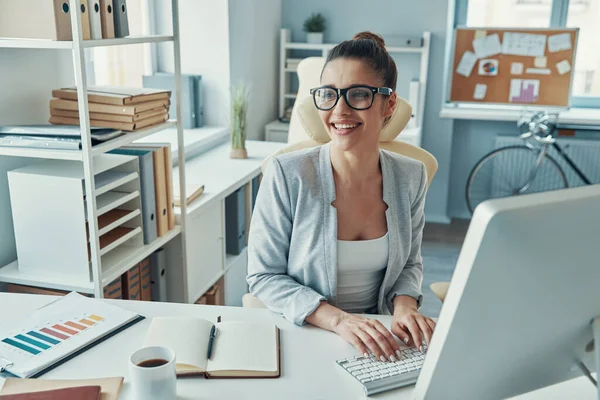 Beautiful young woman in smart casual wear using computer and smiling while working in the office — Φωτογραφία Αρχείου