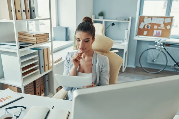 Красива молода жінка в розумному повсякденному одязі, що працює з цифровим планшетом і посміхається, сидячи в офісі — стокове фото