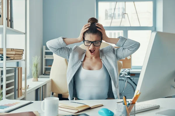 Stressed young woman in smart casual wear keeping head in hands and shouting while sitting in the office — 图库照片