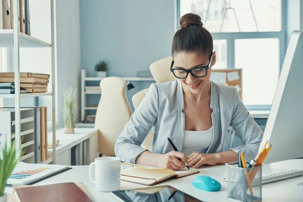 Confident young woman in smart casual wear writing something and smiling while sitting in the office — Photo