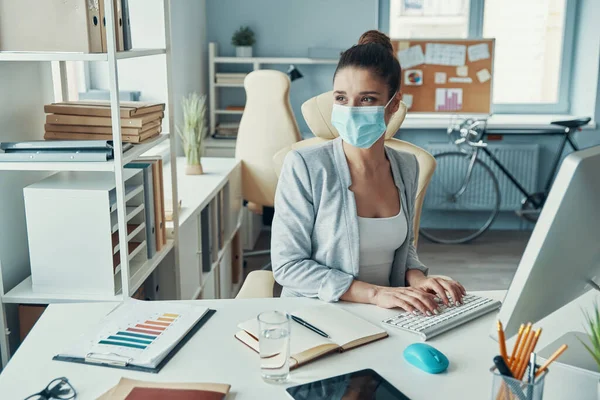 Beautiful young woman in smart casual wear and protective mask using computer — Stock Photo, Image