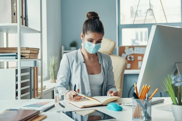 Beautiful young woman in smart casual wear writing something down while sitting in the office — Stock Photo, Image