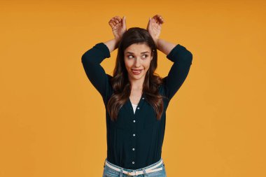 Playful young woman in casual clothing making a face while standing against yellow background