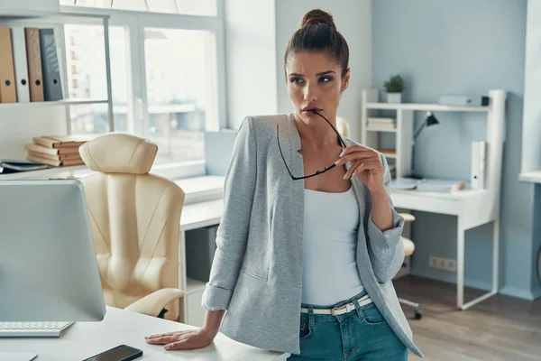 Thoughtful young woman in smart casual looking away and holding eyewear while standing in the office — Stok Foto