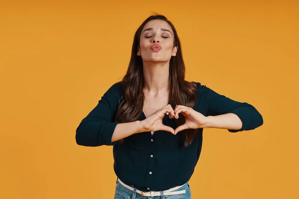 Attractive young woman in casual clothing making heart gesture while standing against yellow background — Fotografia de Stock