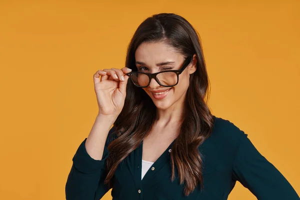 Beautiful young woman in casual clothing smiling and winking while standing against yellow background — Stok fotoğraf