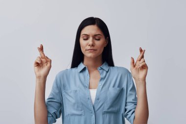 Çekici genç kadın gözlerini kapalı tutuyor ve gri arka planda dururken parmaklarını çapraz yapıyor.