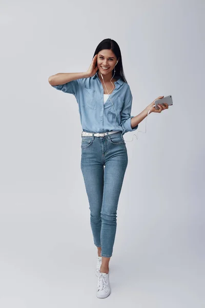 Longitud completa de atractiva joven mujer escuchando música y sonriendo mientras está de pie sobre fondo gris — Foto de Stock