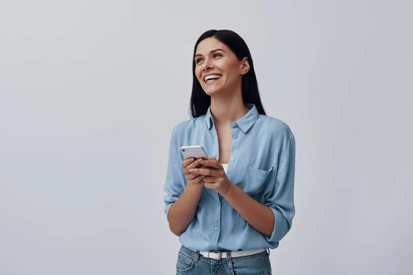 Mujer joven atractiva usando el teléfono inteligente y sonriendo mientras está de pie sobre fondo gris — Foto de Stock