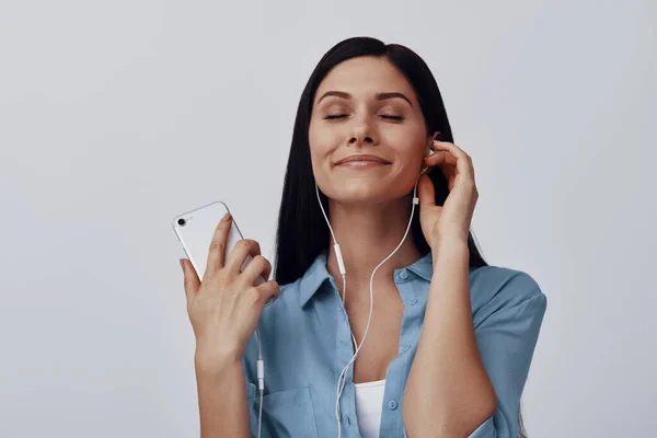 Attraktiv ung kvinna lyssnar musik och ler när du står mot grå bakgrund — Stockfoto
