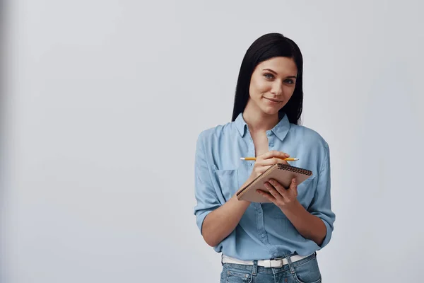 Atractiva joven mujer sosteniendo bloc de notas y mirando a la cámara con sonrisa mientras está de pie sobre un fondo gris — Foto de Stock