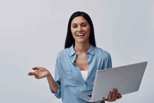 Attraktive junge Frau mit Laptop und lächelndem Blick in die Kamera, während sie vor grauem Hintergrund steht — Stockfoto