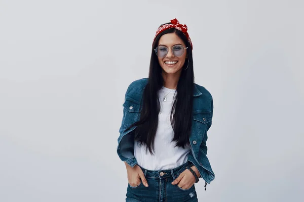 Atractiva joven en bandana mirando a la cámara y sonriendo mientras está de pie sobre un fondo gris —  Fotos de Stock