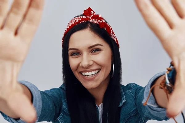 Attraktive junge lächelnde Frau mit Kopftuch blickt in die Kamera und fertigt Handrahmen — Stockfoto