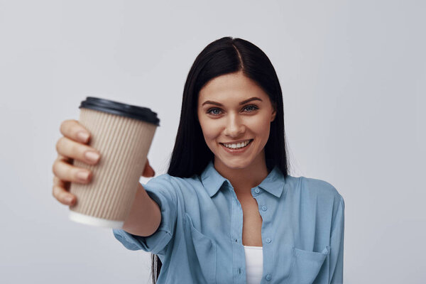Attractive young woman holding disposable cup and smiling while standing against grey background