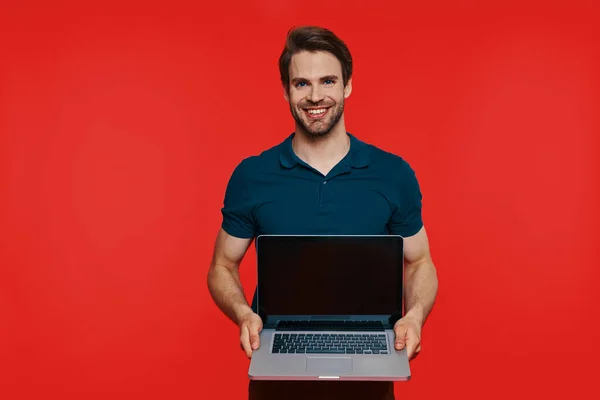 Beau jeune homme souriant en tenue décontractée portant un ordinateur portable et regardant la caméra — Photo