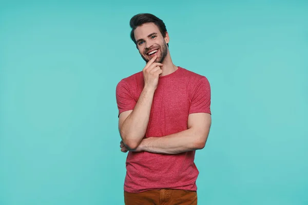 Bonito jovem sorrindo homem em roupas casuais olhando para a câmera e segurando a mão no queixo — Fotografia de Stock